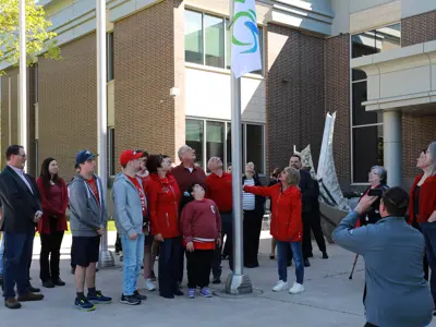 A crowd wearing red shirts looks up as an Accessibility flag is raised atop a pole outside the County of Essex Administration Building. 