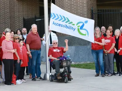 People, many wearing red, hoist an Accessibility flag outside the County of Essex Administration Building.